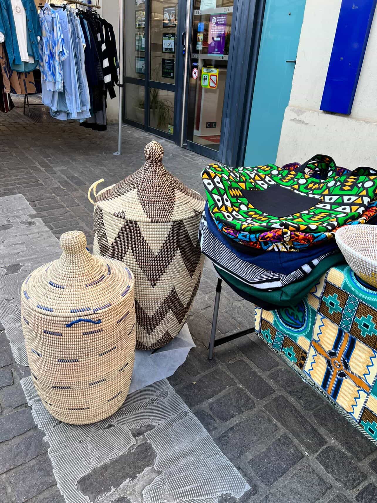 Deux paniers tressés en osier, l'un à motifs zigzag et l'autre uni, sont exposés à côté de vêtements suspendus dans une rue pavée.