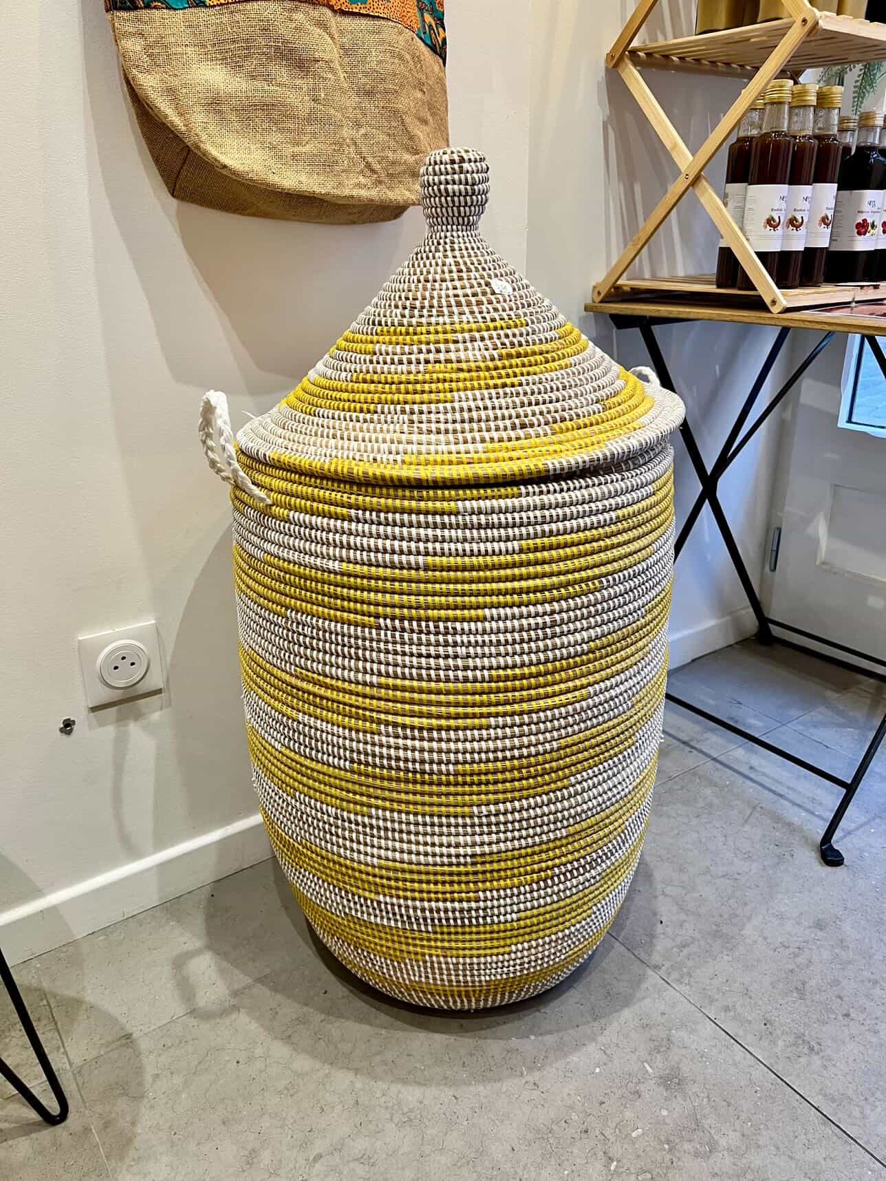 Un grand panier en osier tressé, avec des rayures blanches, jaunes et gris, de forme conique avec un couvercle.