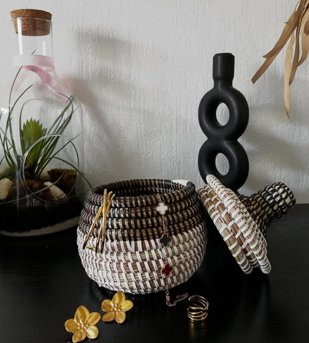 Un pot en osier avec le couvercle ouvert, des accessoires de bijoux à l'intérieur, une plante en pot dans un récipient en verre à côté, et un vase noir à la forme moderne en arrière-plan.