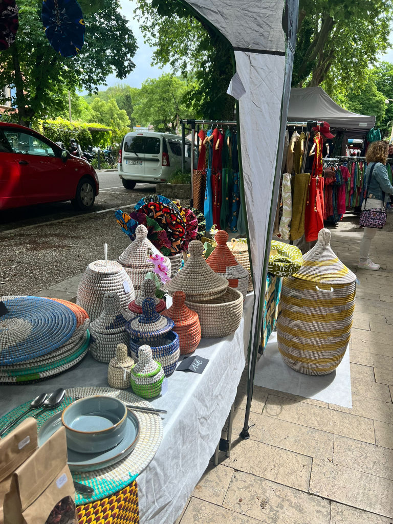 Un étal de marché avec des objets artisanaux, notamment des paniers tressés colorés, des assiettes et des plats, sous un auvent. En arrière-plan, des vêtements colorés sont suspendus sur un portant, avec des voitures stationnées le long de la rue.