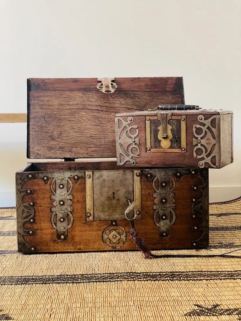 Deux malles anciennes en bois, l'une sur l'autre, ornées de détails métalliques décoratifs, posées sur un tapis en jute.